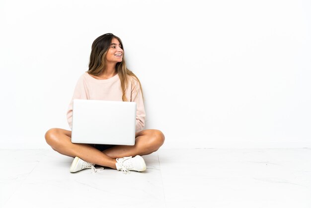 Junge Frau mit einem Laptop, der auf dem Boden lokalisiert auf weißem Hintergrund in seitlicher Position sitzt