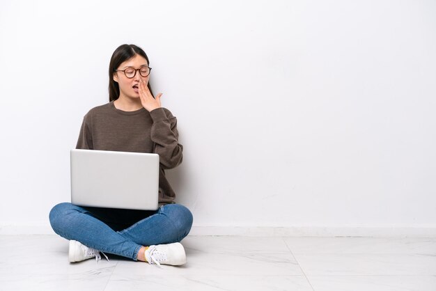 Junge Frau mit einem Laptop, der auf dem Boden sitzt, der gähnt und weit geöffneten Mund mit Hand bedeckt