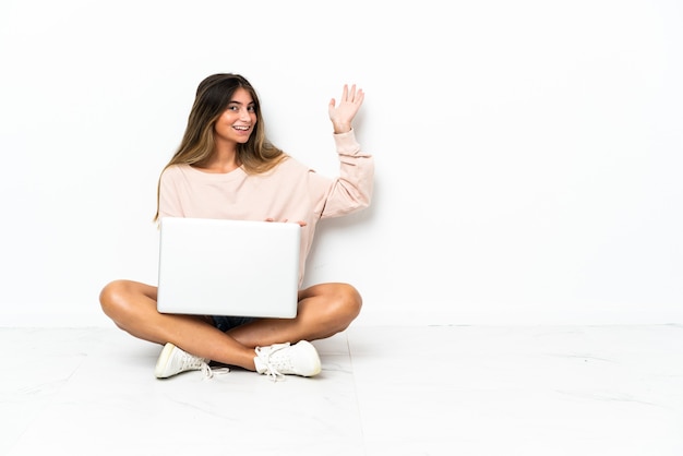 Junge Frau mit einem Laptop, der auf dem Boden sitzt, lokalisiert auf weißer Wand, die Hände zur Seite für Einladung zum Kommen ausdehnt