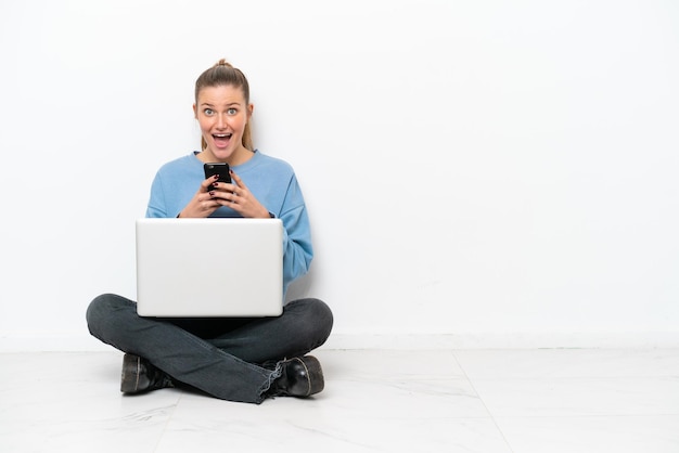 Junge Frau mit einem Laptop, der auf dem Boden sitzt, überrascht und eine Nachricht sendend