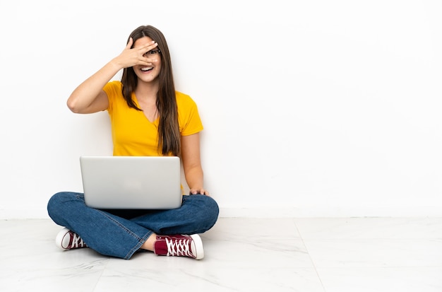 Junge Frau mit einem Laptop, der auf dem Boden sitzt und die Augen mit den Händen bedeckt und lächelt