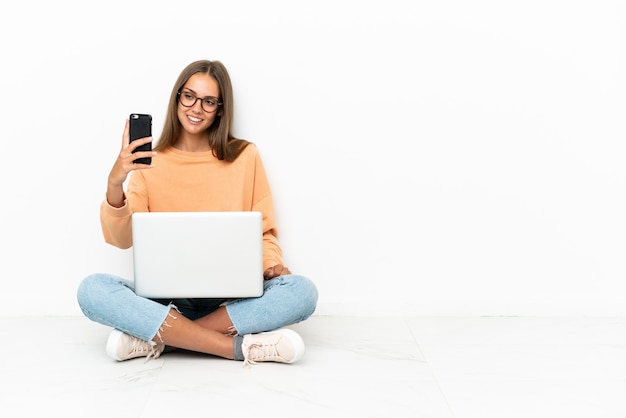 Junge Frau mit einem Laptop, der auf dem Boden sitzt und ein Selfie macht