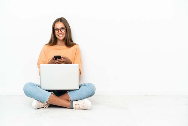 Junge Frau mit einem Laptop, der auf dem Boden sitzt und eine Nachricht mit dem Handy sendet