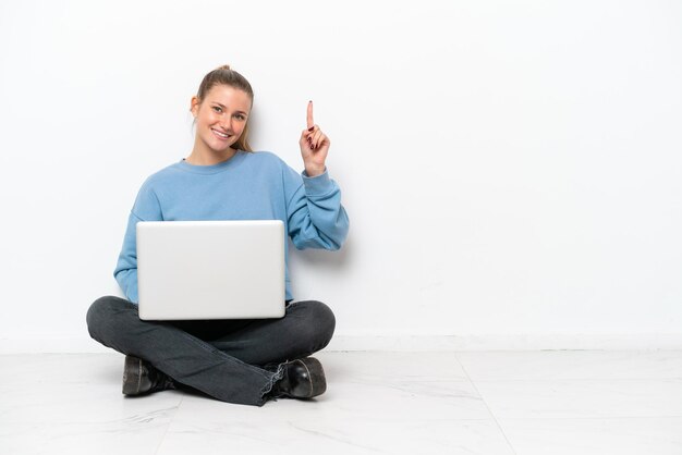 Junge Frau mit einem Laptop, der auf dem Boden sitzt und einen Finger im Zeichen des Besten zeigt und hebt