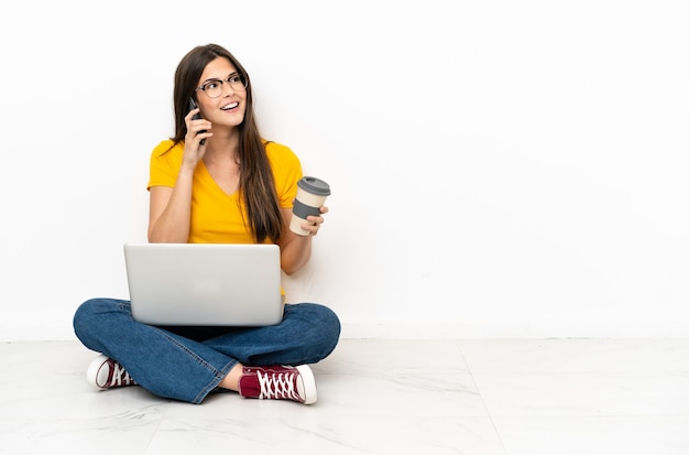 Junge Frau mit einem Laptop, der auf dem Boden sitzt und Kaffee zum Mitnehmen und ein Handy hält