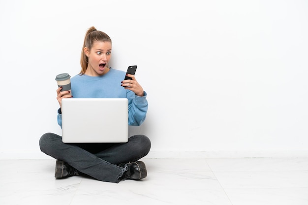 Junge Frau mit einem Laptop, der auf dem Boden sitzt und Kaffee zum Mitnehmen und ein Handy hält