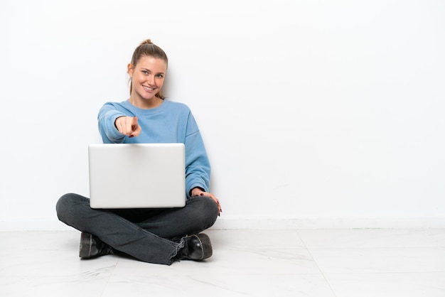 Junge Frau mit einem Laptop, der auf dem Boden sitzt, zeigt Finger mit einem selbstbewussten Ausdruck auf Sie