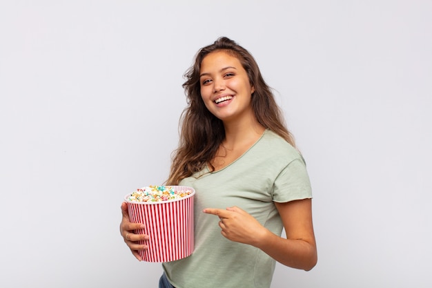 Junge Frau mit einem Pop conrs Eimer, der aufgeregt und überrascht aussieht und zur Seite und nach oben zeigt, um Raum zu kopieren