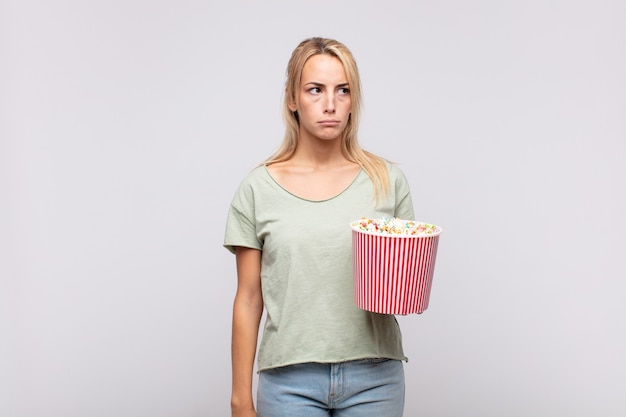 Junge Frau mit einem Popcorn-Eimer, der sich traurig, verärgert oder wütend fühlt und mit einer negativen Einstellung zur Seite schaut und in Uneinigkeit die Stirn runzelt