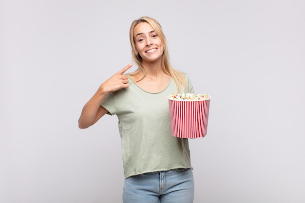 Junge Frau mit einem Popcorn-Eimer lächelt selbstbewusst und zeigt auf ein breites Lächeln, positive, entspannte, zufriedene Haltung