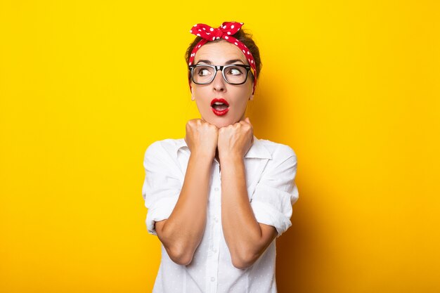 Junge Frau mit einem überraschten Gesicht, Brille und einem Stirnband auf dem Kopf an einer gelben Wand tragend.