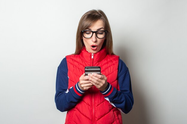 Junge Frau mit einem überraschten Gesicht in einer roten Weste schaut ihr Telefon gegen eine helle Wand an