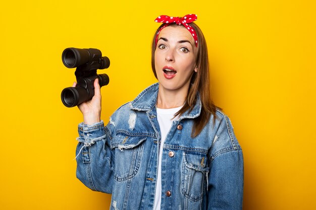 Junge Frau mit einem überraschten Gesicht in Jeans und einem Floß auf dem Kopf hält Fernglas in ihren Händen an einer gelben Wand. Banner.