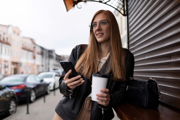 Junge Frau mit einer Tasse Kaffee an einem Mobiltelefon mit einem Lächeln blickt in die Ferne gegen