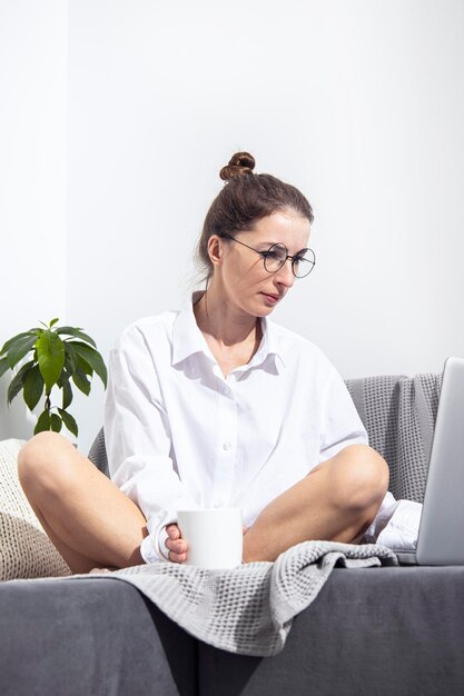 Junge Frau mit einer Tasse Kaffee mit einem Laptop sitzt auf einem Sofa