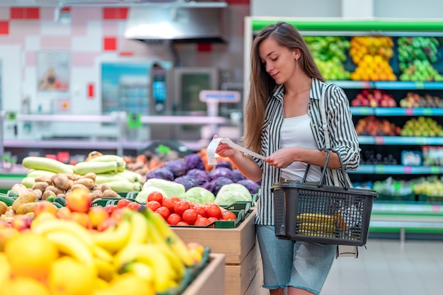 Junge Frau mit Einkaufskorb prüft einen Kaufbeleg nach dem Kauf von Lebensmitteln in einem Supermarkt