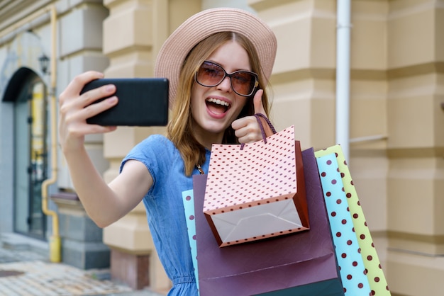 Junge Frau mit Einkaufstaschen machen Selfie, das in einer Stadt am Sommertag geht