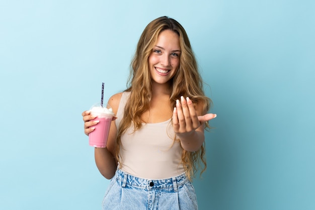 Junge Frau mit Erdbeermilchshake lokalisiert auf Blau, das einlädt, mit Hand zu kommen. Schön, dass du gekommen bist