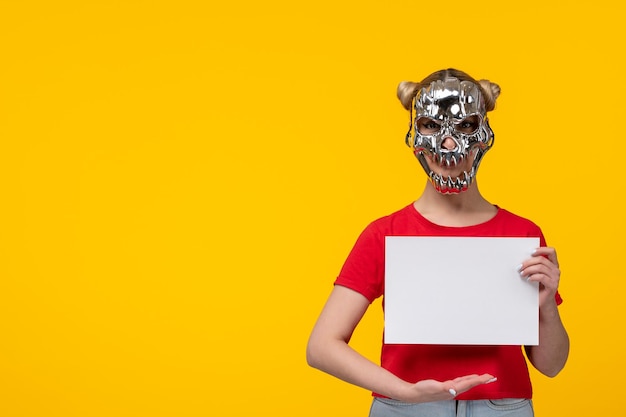 Junge Frau mit erschreckender goldener Maske, die ein Stück Papier auf gelbem Hintergrund hält