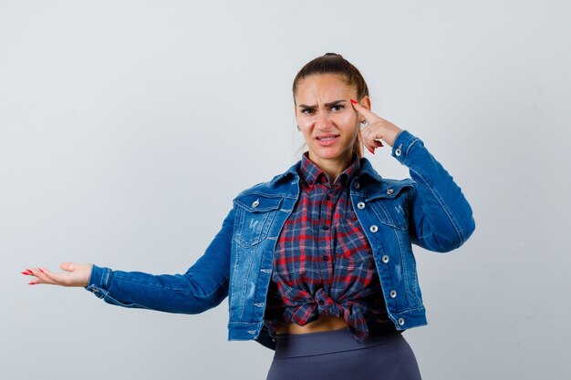 Junge Frau mit Finger an den Schläfen, Arm in kariertem Hemd, Jeansjacke ausbreitend und düster aussehend, Vorderansicht.