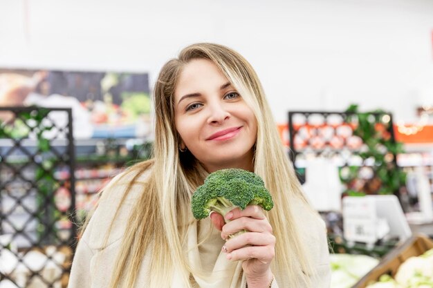 Junge Frau mit frischem Brokkoli im Lebensmittelgeschäft. Lächelnde Blondine in einem beigen Mantel. Gesundes Essen, Vitamine und Vegetarismus. Nahansicht.