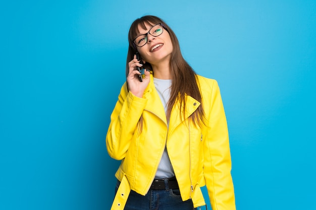 Junge Frau mit gelber Jacke auf dem blauen Hintergrund, der ein Gespräch mit dem Handy hält