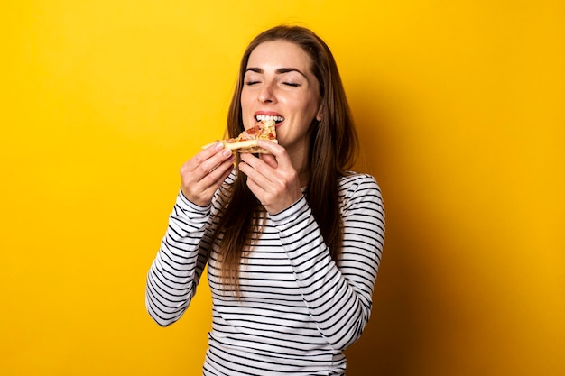 Junge Frau mit geschlossenen Augen, die ein Stück heiße frische Pizza auf gelbem Hintergrund isst