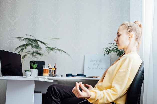 Junge Frau mit geschlossenen Augen in der Nähe des Laptops im Heimbüro, die Meditation am Tisch praktiziert, Freiberuflerin