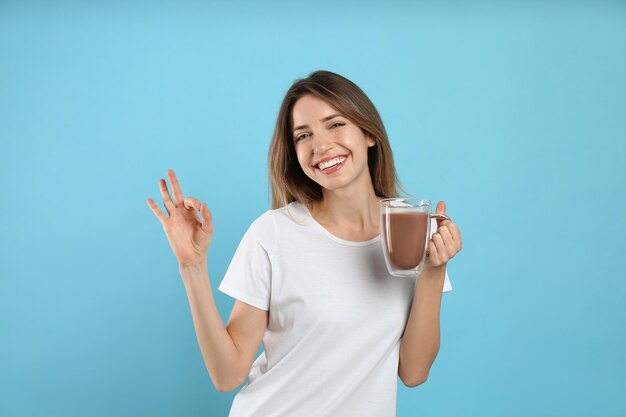 Junge Frau mit Glastasse Schokoladenmilch, die Ok auf hellblauem Hintergrund zeigt