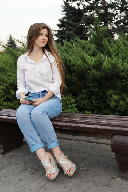 Junge Frau mit glatten langen Haaren allein sitzt friedlich auf der Parkbank und hält Smartphone