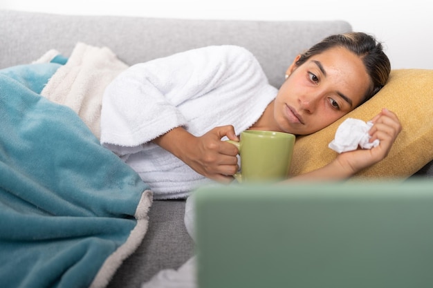 Junge Frau mit Grippe, die eine Tasse grünen Tee hält und einen Film auf einem digitalen Tablet ansieht