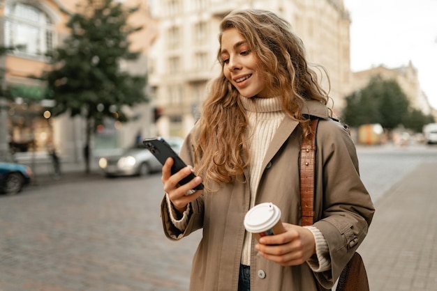 Junge Frau mit Herbstmantel, die mit Smartphone und Kaffeetasse in einer Stadtstraße spazieren geht