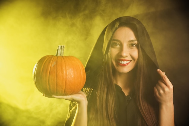 Junge Frau mit Hexenkostüm mit Kürbis in Rauchwolke auf dunklem Hintergrund Halloween-Party