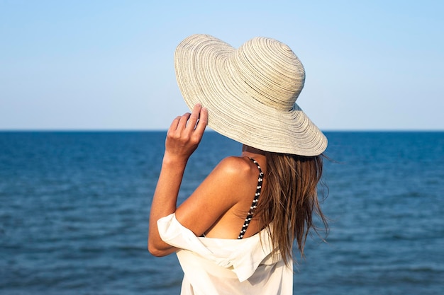 Junge Frau mit Hut auf einer Klippe blickt an einem Sommertag auf das Meer.