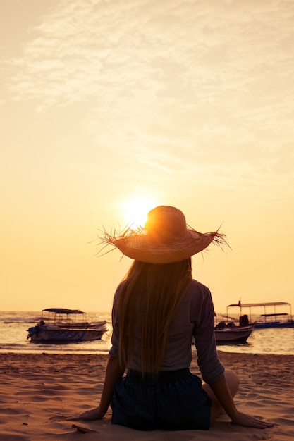 Foto junge frau mit hut, die den sonnenuntergang im ozean betrachtet