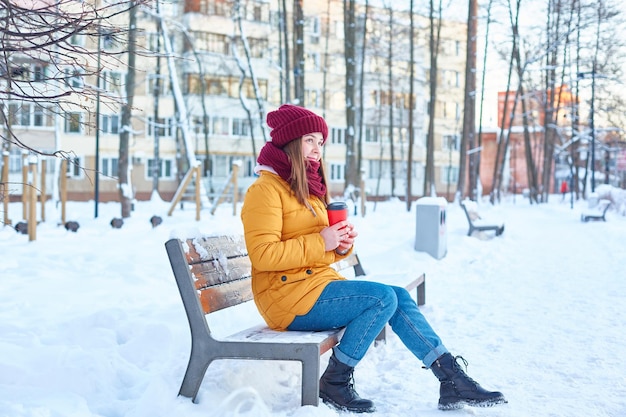 junge Frau mit Hut und gelber Jacke mit einer Einwegtasse Kaffee in den Händen an einem frostigen Wintertag auf einer Bank