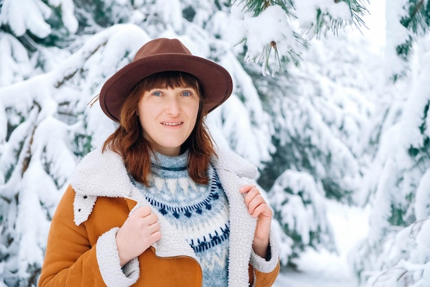 Junge Frau mit Hut und warmer Jacke, die zwischen schneebedeckten Bäumen steht und den ersten Schnee genießt