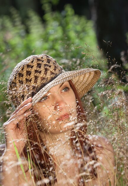 Junge Frau mit Hut unter dem Gras, das von den Sonnenstrahlen auf einem Waldhintergrund beleuchtet wird