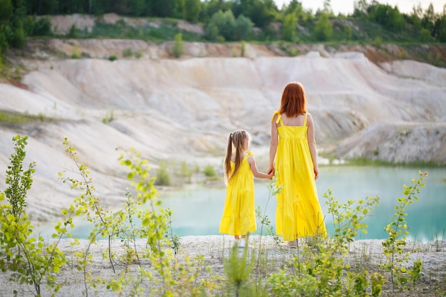 Junge Frau mit ihrer Tochter in einem gelben Kleid nahe dem See mit azurblauem Wasser und grünen Bäumen. Glückliches Familienbeziehungskonzept