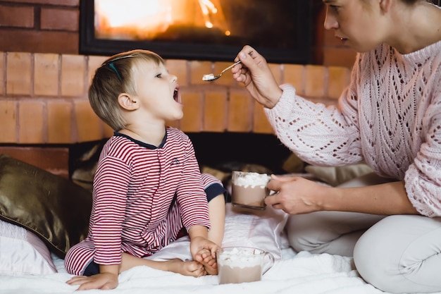 Foto junge frau mit kind am kamin. mutter und sohn trinken kakao mit marshmello am kamin.