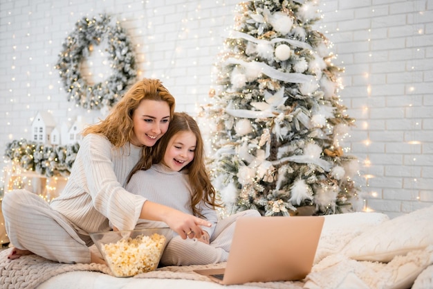 Junge Frau mit Kind, Baby, 7 Jahre alt, in Nachtwäsche, die Filme anschaut und Popcorn auf dem Bett in weihnachtlich dekoriertem Zuhause isst