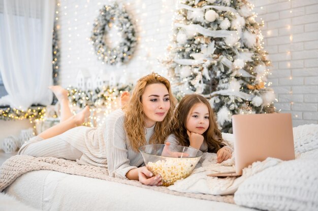 Junge Frau mit Kind, Baby, 7 Jahre alt, in Nachtwäsche, die Filme anschaut und Popcorn auf dem Bett in weihnachtlich dekoriertem Zuhause isst