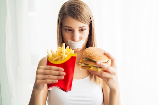 Junge Frau mit Klebeband über dem Mund, die sie daran hindert, Junk Food zu essen.