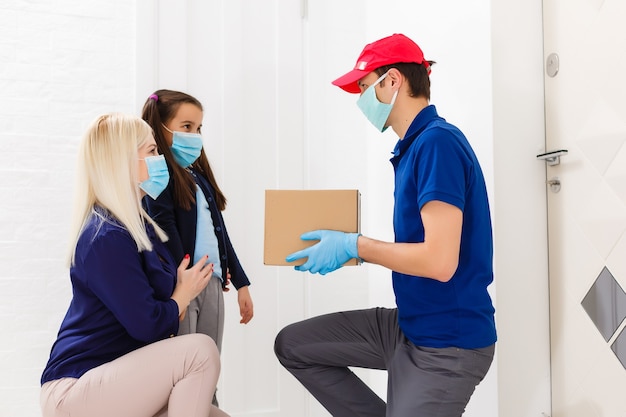 Junge Frau mit kleiner Tochter mit medizinischer Maske, die bestelltes Essen vom Liefermann im Haus erhält. Verhinderung der Virusausbreitung