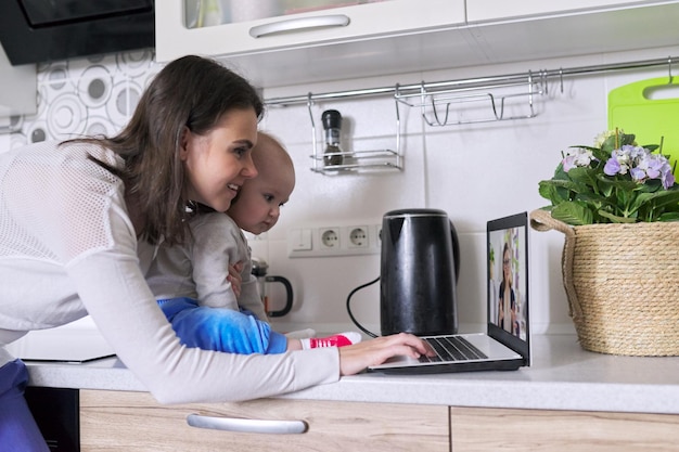 Junge Frau mit Kleinkind in den Armen, die Videoanrufe macht, Videokonferenz mit Laptop zu Hause. Remote-Arbeit, Online-Kommunikation mit Kollegen, Kunden. Technologie, Distanz-Lifestyle-Konzept