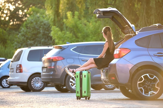 Junge Frau mit Koffertasche, die in der Nähe ihres Autos wartet Reise- und Urlaubskonzept