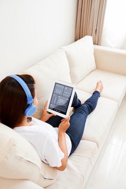 Junge Frau mit Kopfhörern, die auf dem Sofa sitzt und die Anwendung auf dem Tablet-Computer verwendet, um die Musiklautstärke einzustellen