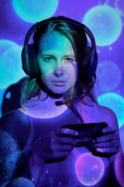 Junge Frau mit Kopfhörern und Controller schaut mit ernsthaftem Gesicht in die Kamera, während sie unter bunter Beleuchtung ein schwieriges Videospiel spielt