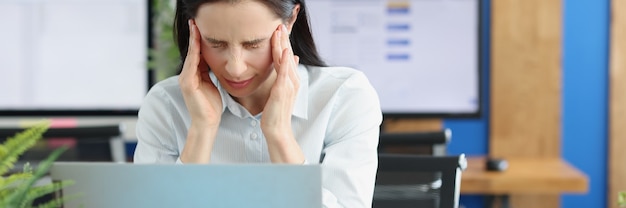 Junge Frau mit Kopfschmerzen hinter Laptop am Arbeitsplatz