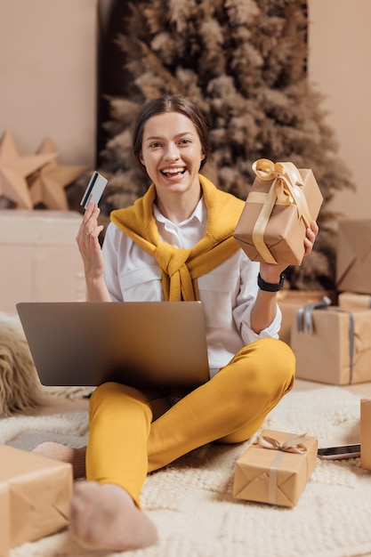 Junge Frau mit Kreditkarte, die online auf Laptop in gemütlichem Weihnachtsinterieur einkauft